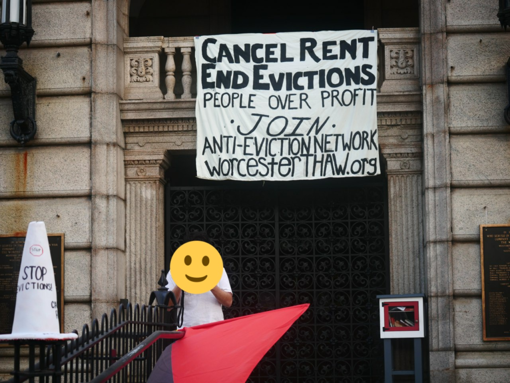 A person stands in front of a stone building with a banner behind them which reads "Cancel Rent, End Evictions. People Over Profit. Join Anti Eviction Network WorcesterThaw.org". The face of the speaker is obscured by a smiley face emoji. 